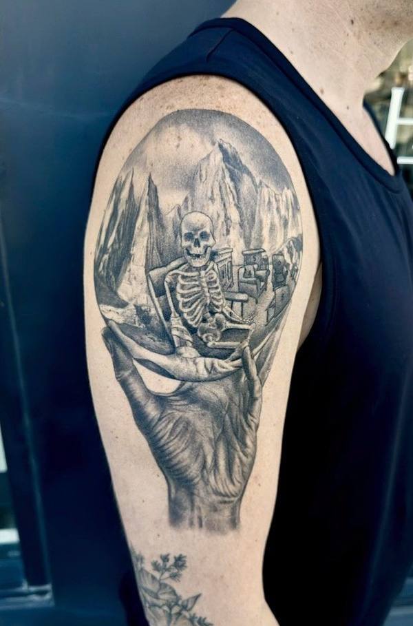 Black and gray tattoo of a skeleton held in a hand with a mountain landscape by Cihan Pohrenk